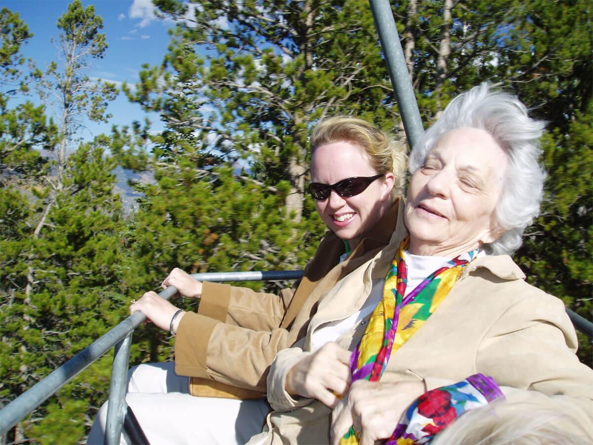 A senior woman and her carer are riding a cable car. They both laugh.